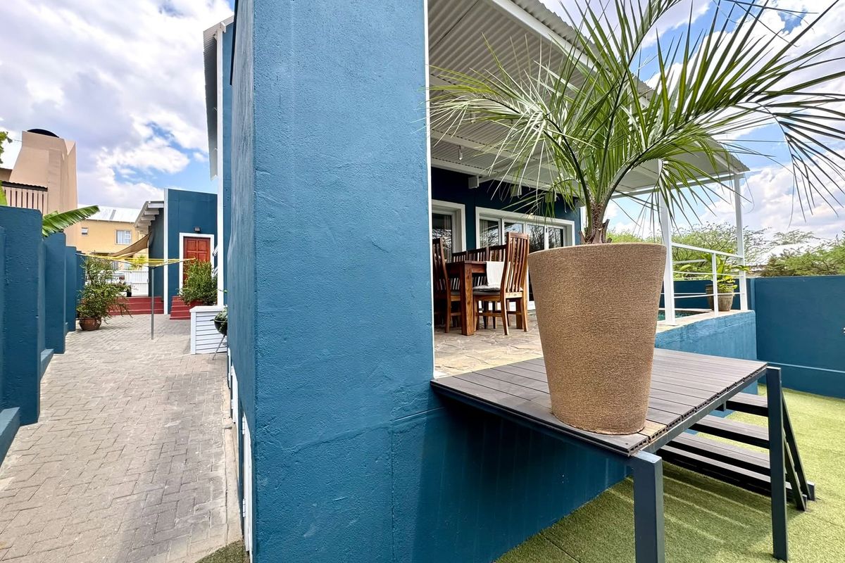 A well-maintained rear exterior area featuring a covered patio, paved pathways, artificial lawn, and a raised deck platform.