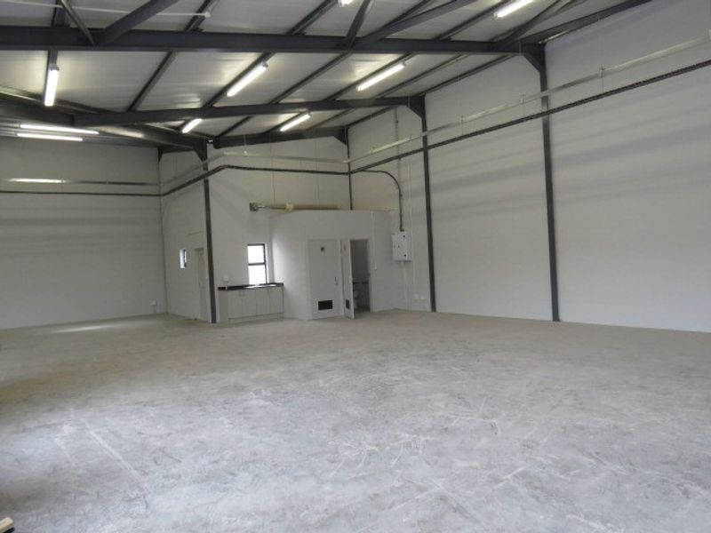 Spacious industrial warehouse unit featuring high ceilings with exposed steel beams, concrete flooring, fluorescent lighting, and a small integrated kitchenette area.