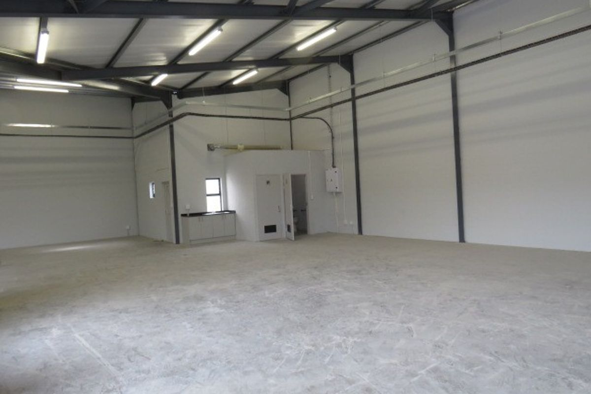 Spacious industrial warehouse unit featuring high ceilings with exposed steel beams, concrete flooring, fluorescent lighting, and a small integrated kitchenette area.