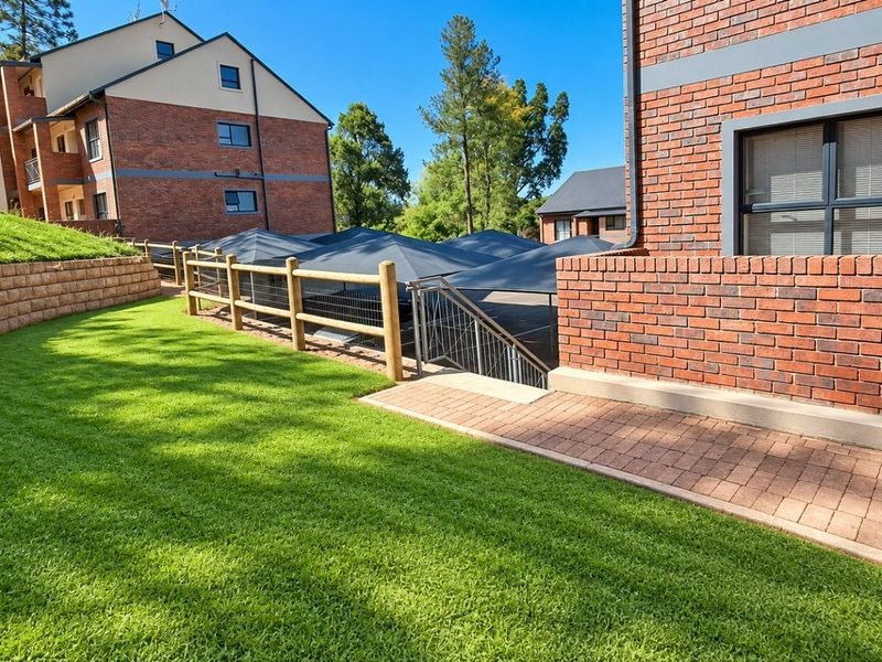 A well-maintained exterior view of the apartment complex featuring brick facades, landscaped lawn, retaining walls, and a paved walkway leading to a shaded parking area.