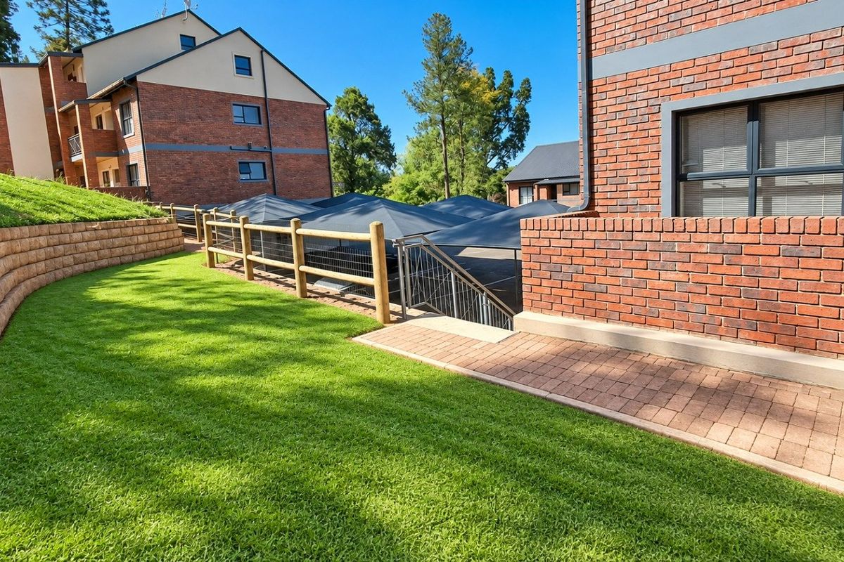 A well-maintained exterior view of the apartment complex featuring brick facades, landscaped lawn, retaining walls, and a paved walkway leading to a shaded parking area.
