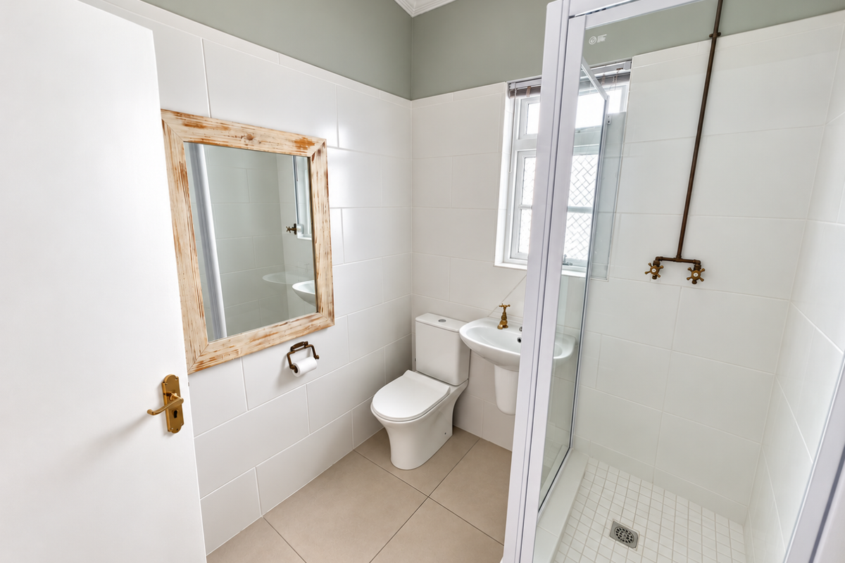 A clean, modern bathroom featuring a glass-enclosed shower, pedestal basin, toilet, and neutral tiled finishes with brass-toned fixtures.