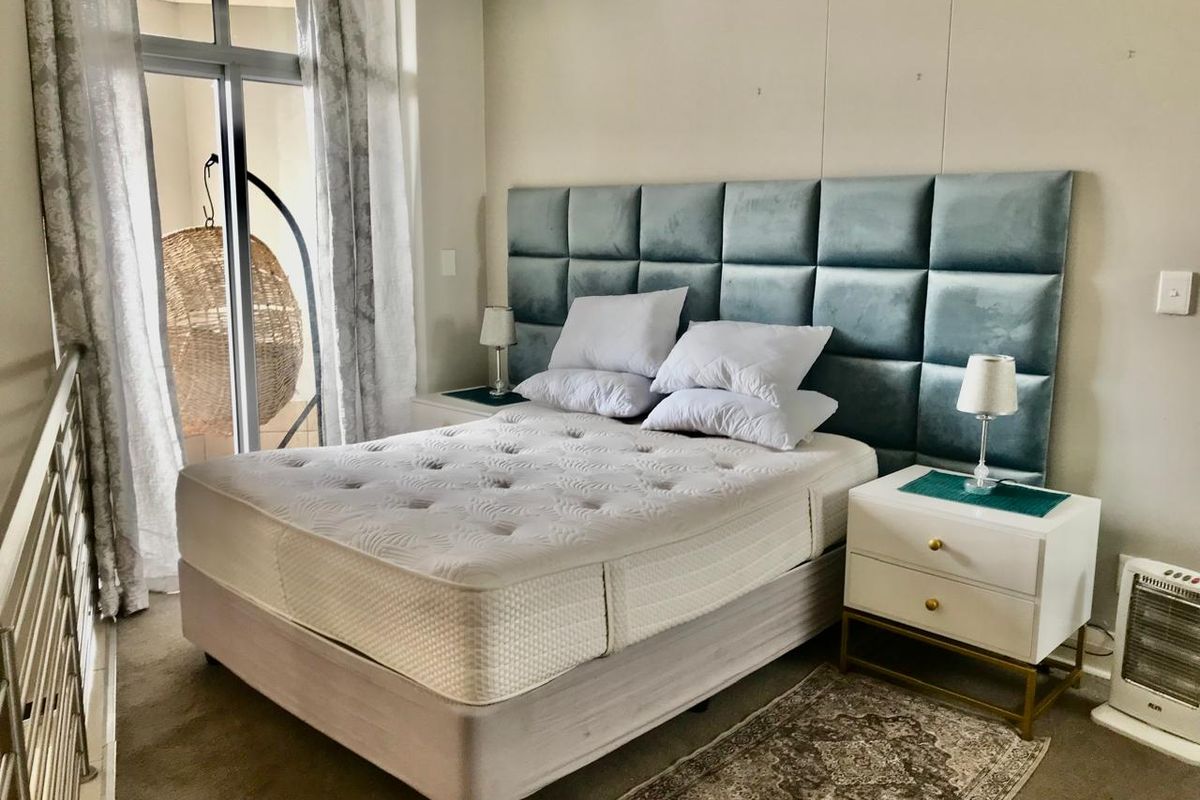 A well-lit bedroom featuring a large upholstered headboard, neutral carpeting, and direct access to a private balcony through sliding glass doors.
