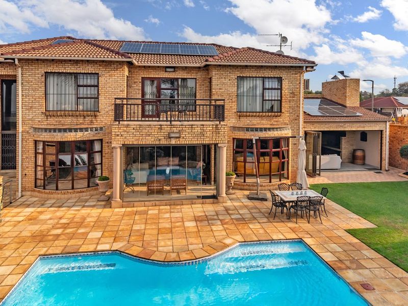 Rear view of a double-story brick house featuring a swimming pool, paved patio, balcony, and integrated solar panels on the tiled roof.