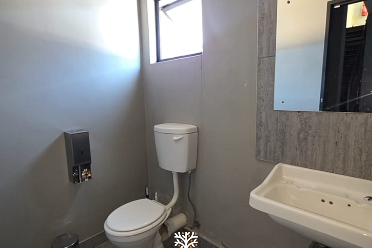 A functional industrial-style restroom featuring a wall-mounted basin, standard toilet, and stainless steel soap dispenser with tiled flooring.