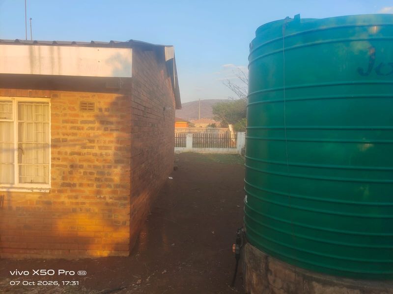 Rear exterior view showing a brick building structure, a large green water storage tank, and a perimeter fence in the background.
