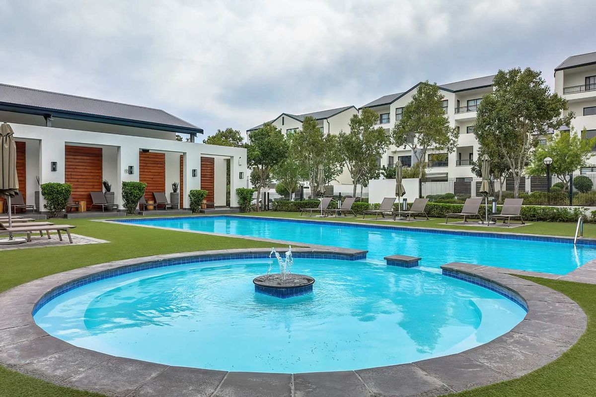 A well-maintained communal swimming pool area featuring a central fountain, tiled coping, artificial turf, and a covered patio structure with timber accents.