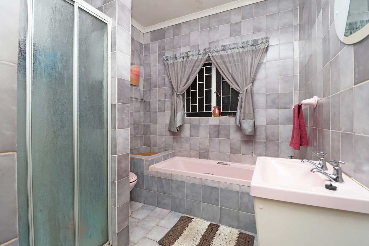 A dated bathroom featuring a pink bathtub and basin, tiled walls, and a separate shower enclosure with security bars on the window.