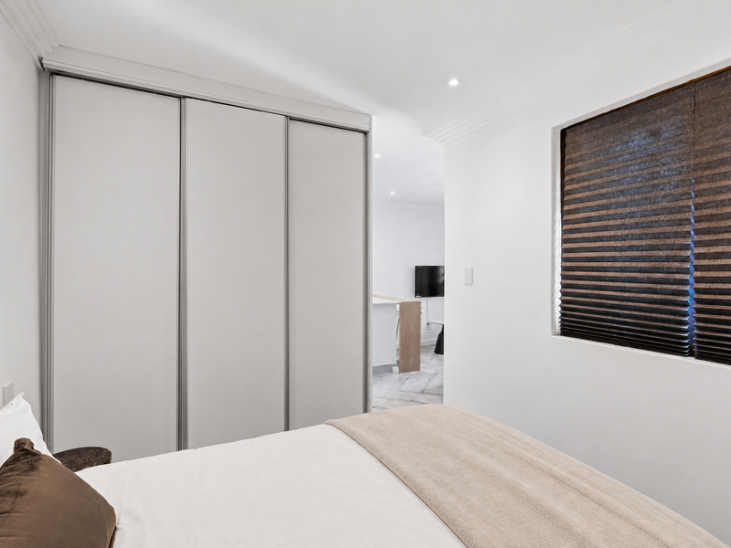 A clean, modern bedroom featuring a large built-in sliding wardrobe, recessed downlighting, and a window with a fitted pleated blind.