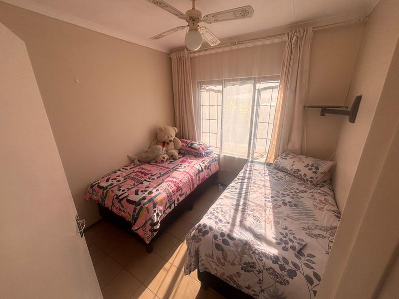 A compact bedroom featuring tiled flooring, a ceiling fan, and a window with security bars and curtain rails.