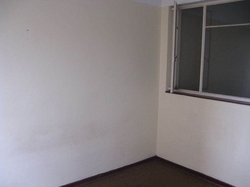 An empty bedroom featuring plain walls, a metal frame window, and dark skirting boards, requiring cosmetic updates to improve its overall condition.
