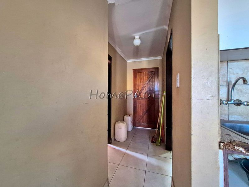 A narrow hallway with tiled flooring leading to a wooden door, showing signs of wear on the walls and adjacent kitchen cabinetry.