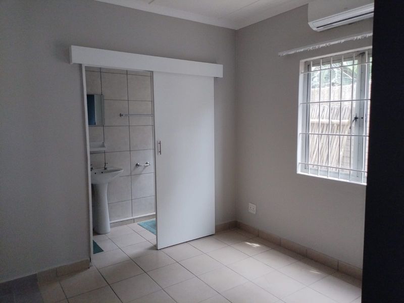 A clean, empty bedroom featuring tiled floors, an air conditioning unit, and an en-suite bathroom accessed via a modern sliding door.