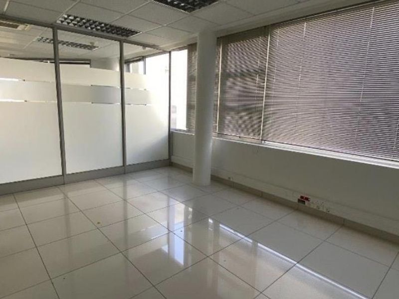 A bright, vacant office space featuring tiled flooring, glass partitions, and large windows with venetian blinds, suitable for a professional workspace.