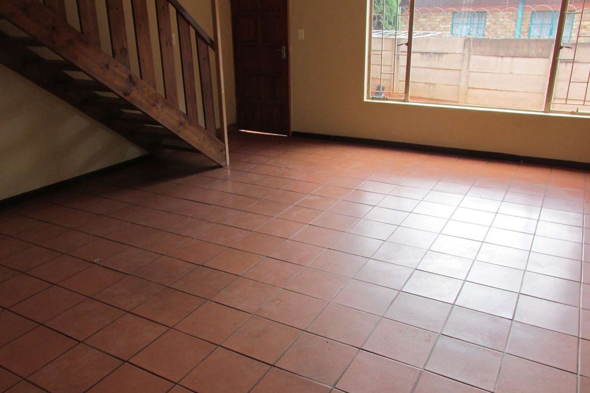 An empty living area featuring terracotta tiled flooring, a wooden staircase, and a large window with security bars.
