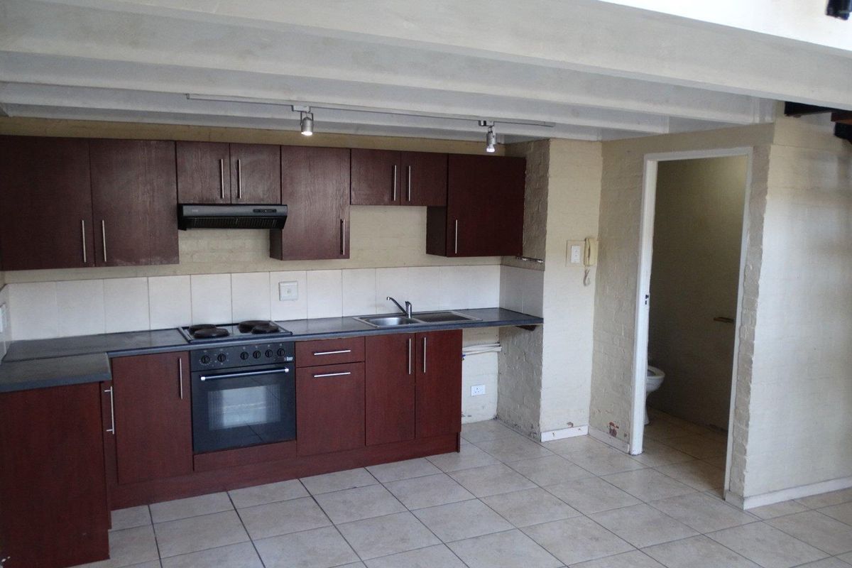 A functional kitchen featuring dark wood cabinetry, built-in oven and hob, tiled flooring, and exposed ceiling beams, with an adjacent bathroom entrance.