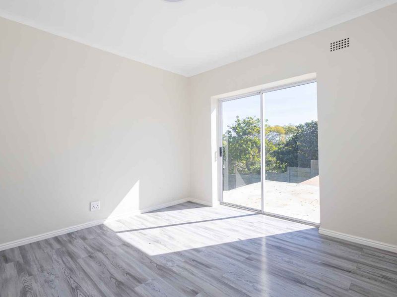 A bright, empty bedroom featuring modern laminate flooring, a built-in cupboard, and a sliding glass door providing access to an outdoor area.