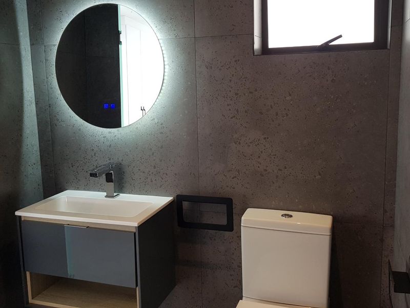 Modern bathroom featuring dark grey tiled walls, a floating vanity with basin, a backlit circular mirror, and a contemporary toilet.