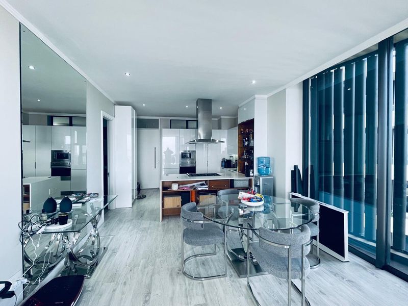 Modern open-plan kitchen and dining area featuring sleek white cabinetry, integrated appliances, recessed lighting, and large windows with vertical blinds.