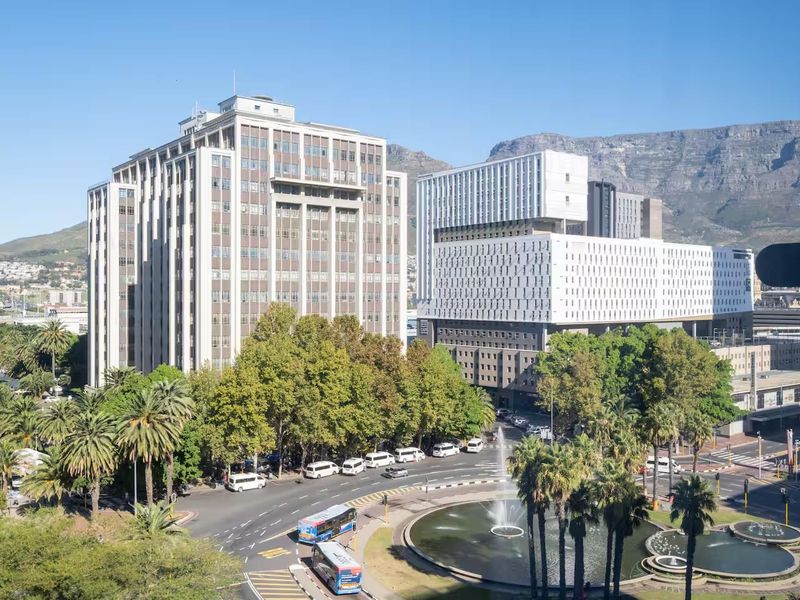 Elevated urban view overlooking a city roundabout, fountain, and surrounding high-rise buildings with mountain scenery in the background.