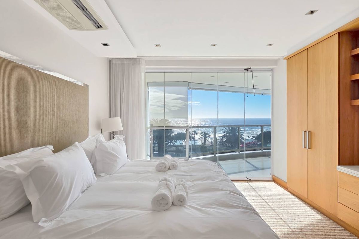 A bright, modern bedroom featuring built-in wooden cabinetry, recessed lighting, and floor-to-ceiling glass doors opening onto a balcony with ocean views.