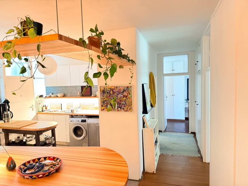 An open-plan kitchen area featuring modern cabinetry, a tiled backsplash, and integrated laundry appliances, flowing into a bright hallway with built-in storage.