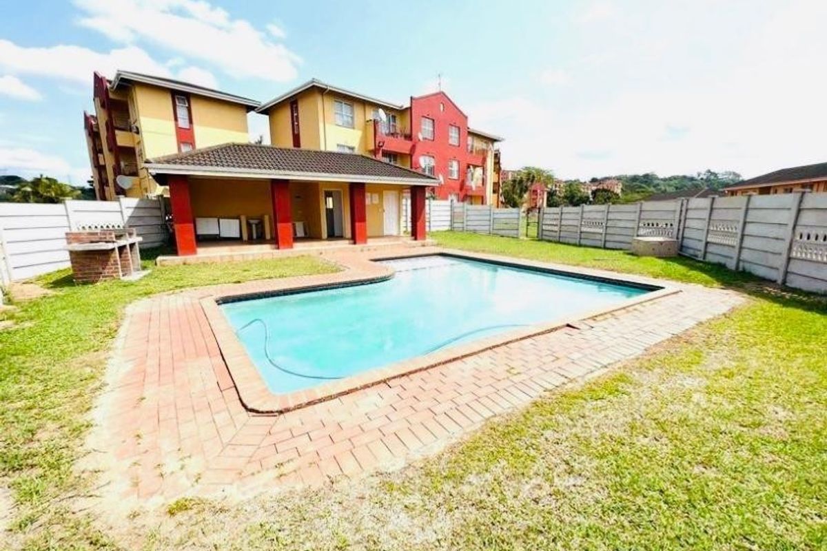 Communal pool area featuring a swimming pool, brick paved surround, covered patio, and built-in braai facility within a fenced complex.