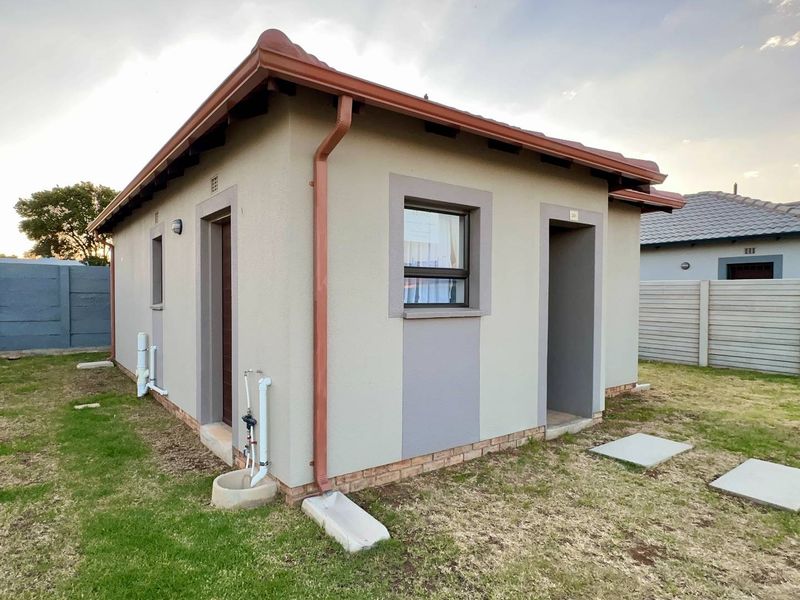 A view of the rear exterior of a small residential unit featuring a tiled roof, external plumbing connections, and a surrounding garden area.