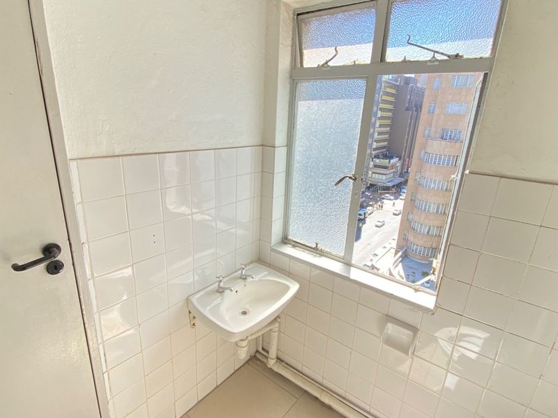 A compact utility area featuring a wall-mounted basin, tiled splashback, and a window overlooking the city street.