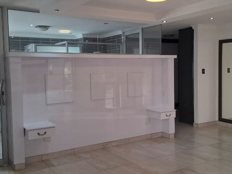 Spacious bedroom featuring a modern glass-enclosed en-suite bathroom, built-in headboard with integrated bedside tables, and neutral tiled flooring throughout.
