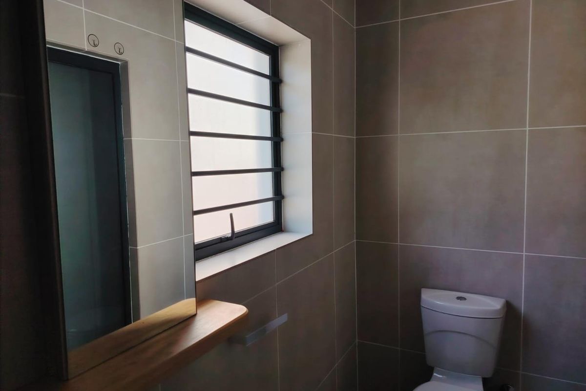 A modern, neutral-toned bathroom featuring floor-to-ceiling tiling, a wall-mounted mirror with a wooden shelf, and a secure window with fitted bars.