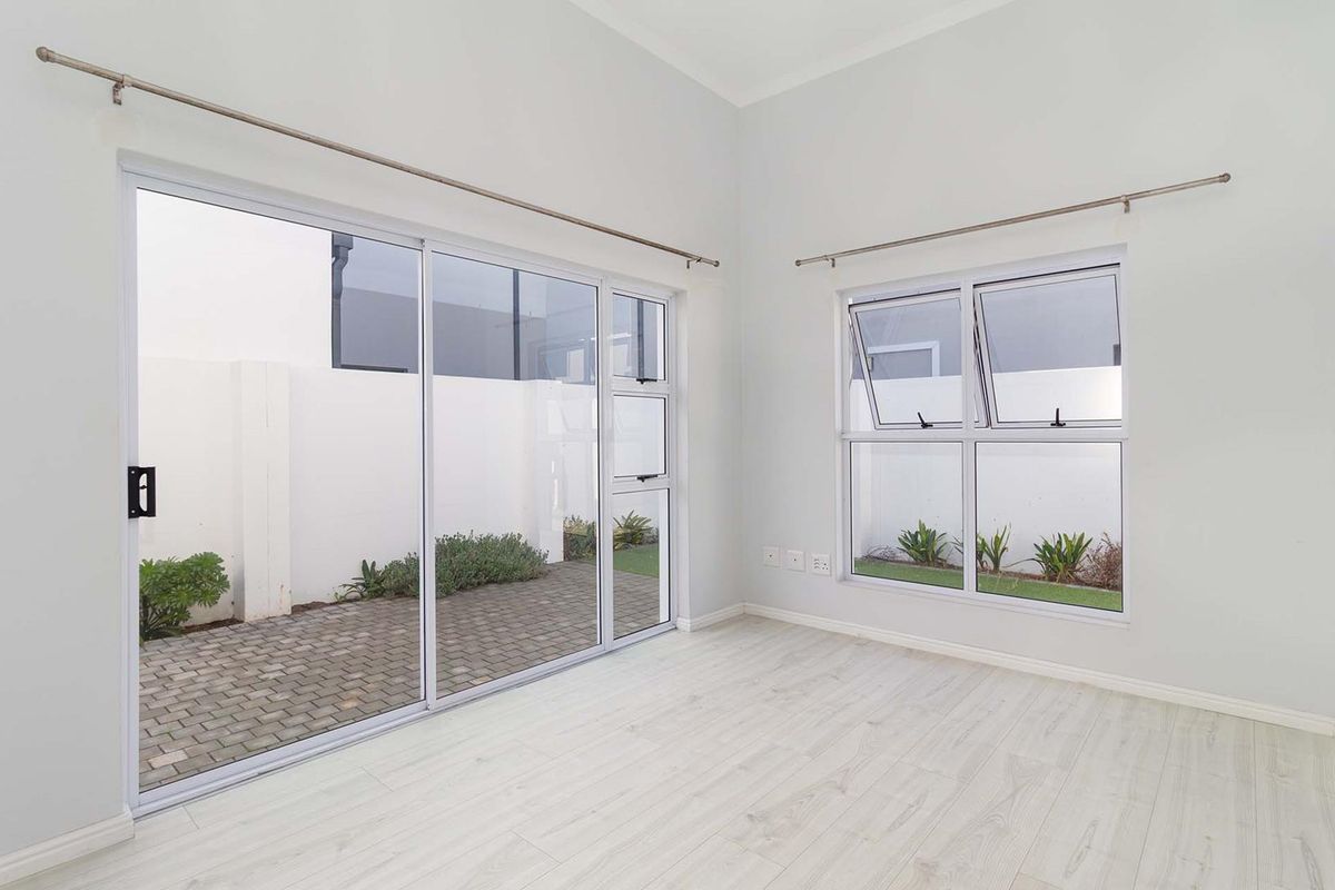 A bright, empty bedroom featuring light-toned laminate flooring, large sliding glass doors leading to a paved patio, and a top-hung window.