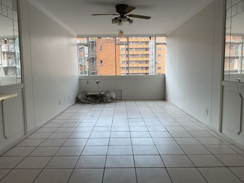 An empty, tiled living room featuring a ceiling fan, large window, and decorative mirrored wall panels, offering a blank canvas for renovation.