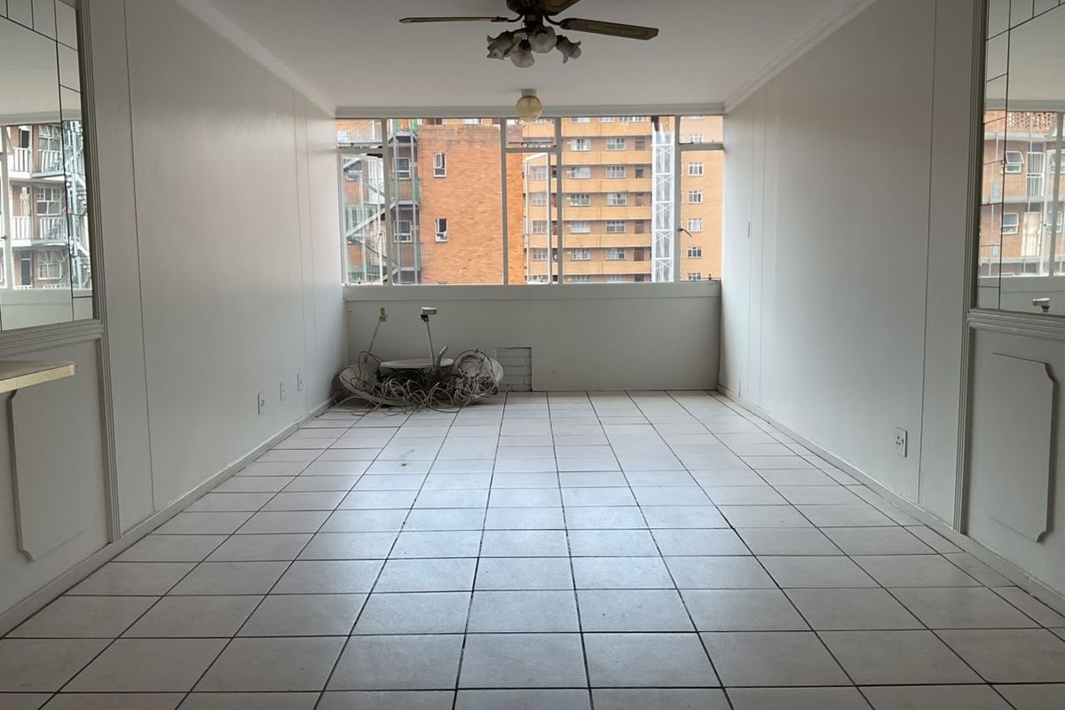 An empty, tiled living room featuring a ceiling fan, large window, and decorative mirrored wall panels, offering a blank canvas for renovation.
