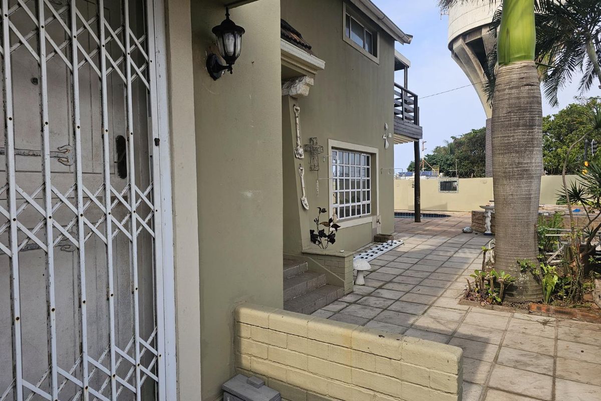 A rear exterior view of the property featuring a paved patio area, security gate, and a balcony overlooking the garden space.