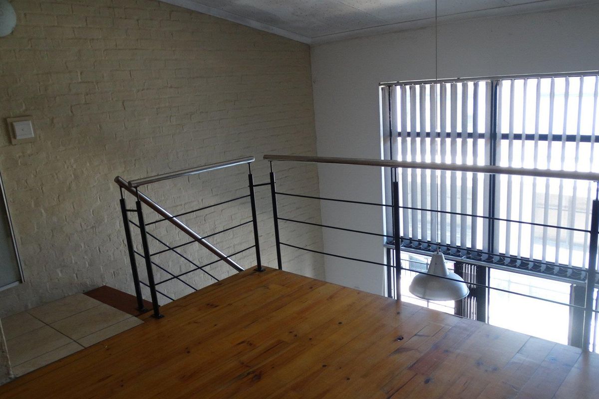 An open mezzanine level featuring timber flooring, a modern stainless steel balustrade, and an exposed brick wall with large windows and vertical blinds.