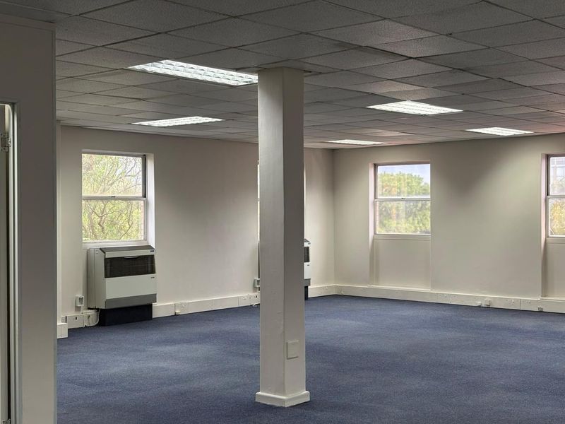 A spacious, empty office area featuring a suspended ceiling, recessed lighting, carpeted flooring, and wall-mounted air conditioning units.