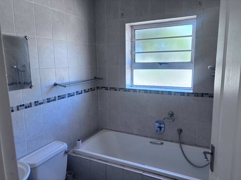 A functional bathroom featuring a bathtub with shower mixer, tiled walls with decorative border, and a window with security bars.