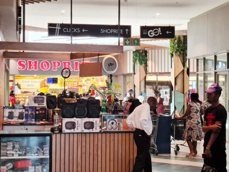 A busy commercial shopping centre corridor featuring glass storefronts, directional signage, and modern tiled flooring with timber-look architectural accents.