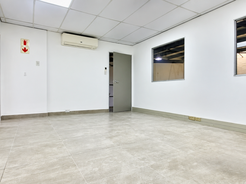 A clean, empty office space featuring tiled flooring, a suspended ceiling, split-system air conditioning, and internal windows looking into the warehouse area.