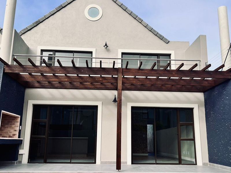 Rear exterior view of a townhouse featuring a tiled patio, timber pergola, built-in braai, and a balcony with stainless steel railings.