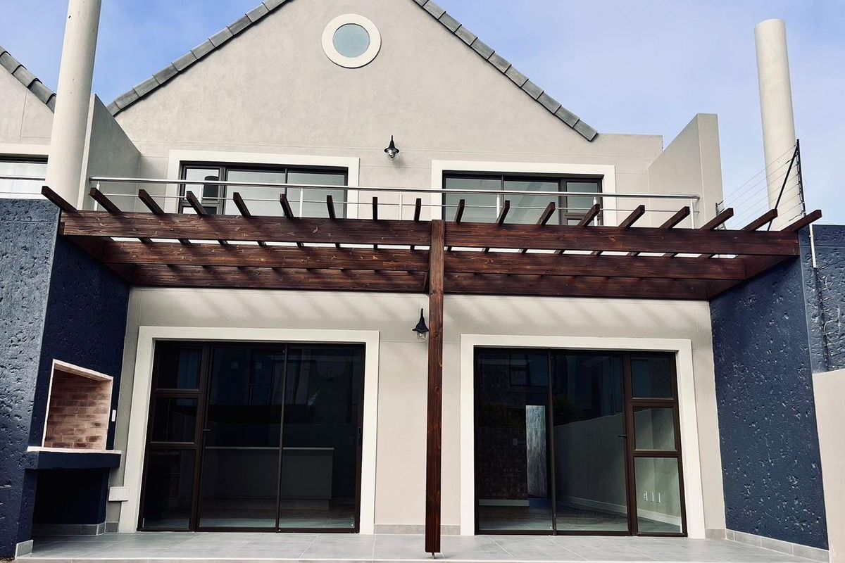 Rear exterior view of a townhouse featuring a tiled patio, timber pergola, built-in braai, and a balcony with stainless steel railings.
