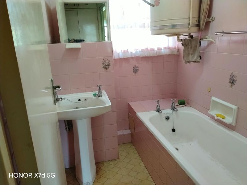 A dated bathroom featuring pink wall tiles, a pedestal basin, a standard bathtub, and a wall-mounted geyser unit.
