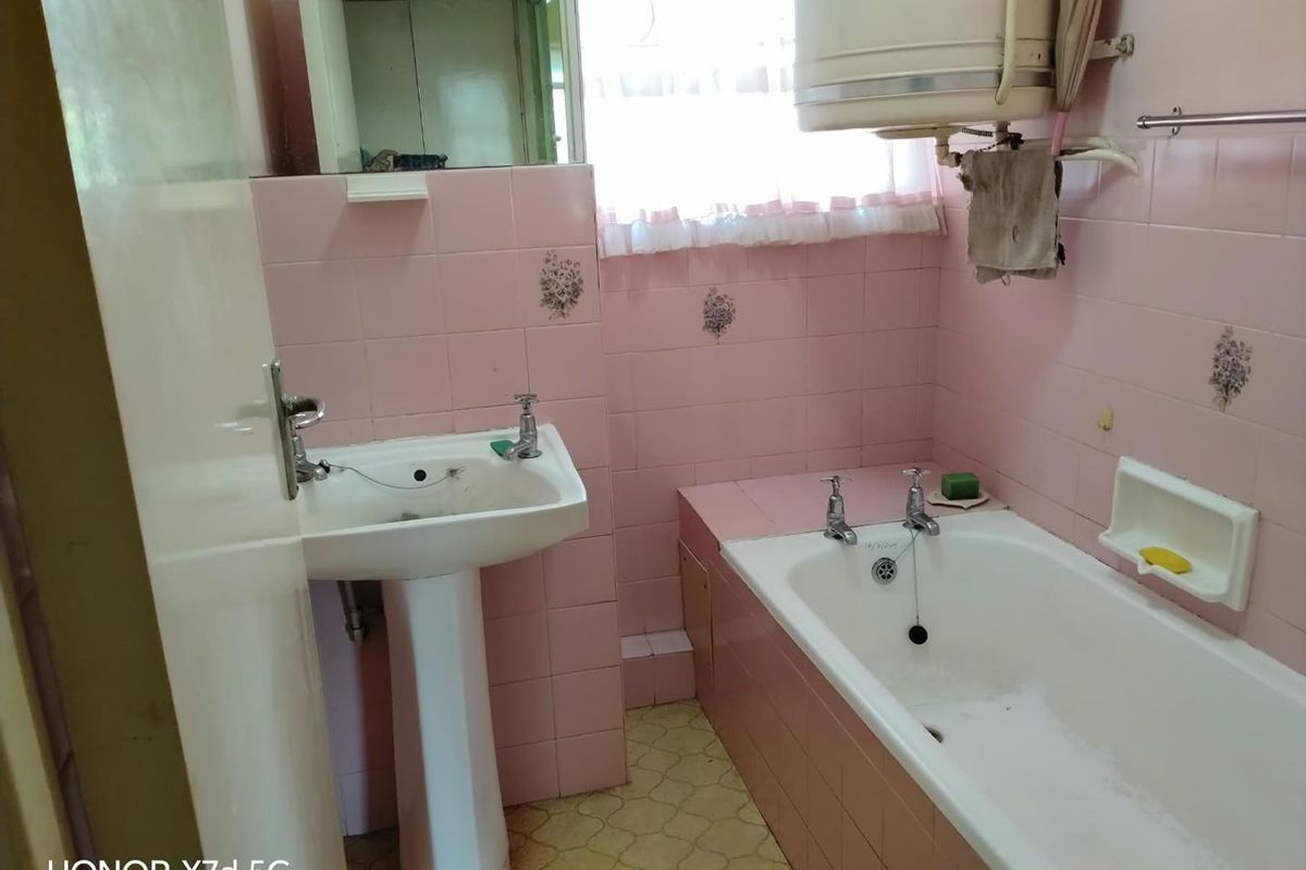A dated bathroom featuring pink wall tiles, a pedestal basin, a standard bathtub, and a wall-mounted geyser unit.