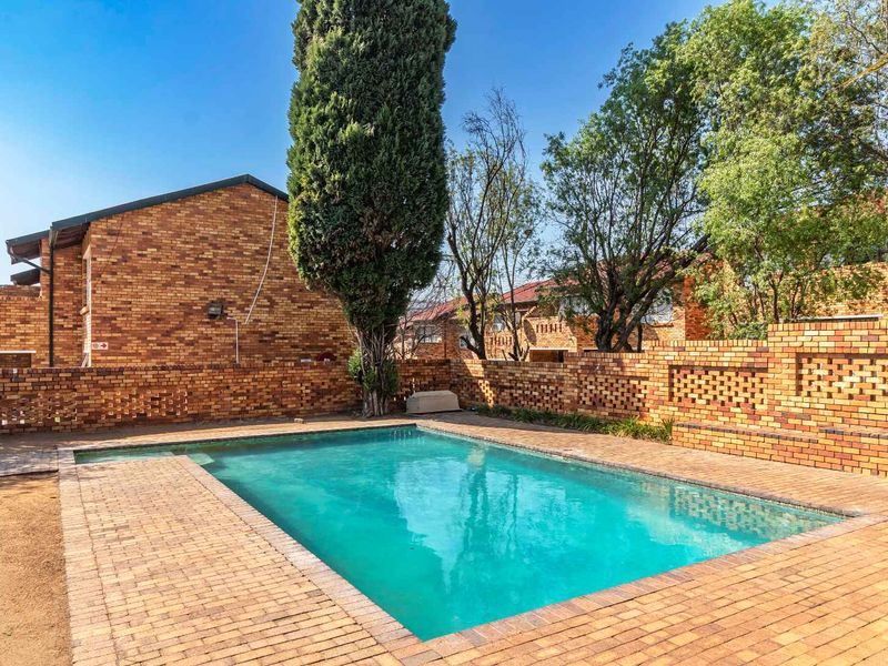 A communal swimming pool area featuring brick paving, a perimeter brick wall, and established trees within an apartment complex setting.