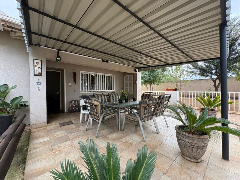 A covered outdoor patio area featuring tiled flooring, a corrugated metal roof, and security bars on the windows, providing a functional space for outdoor living.
