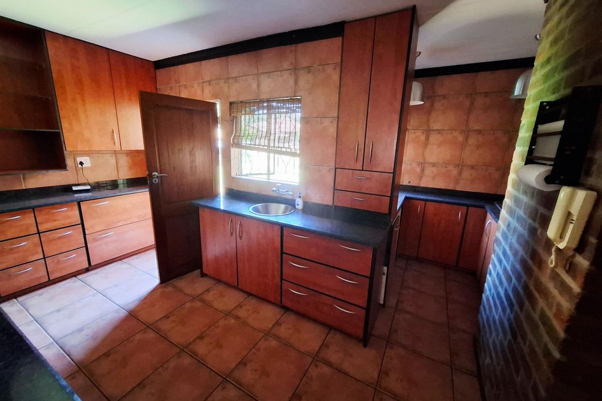 A functional kitchen featuring wooden cabinetry, dark stone countertops, tiled splashbacks, and tiled flooring, with an adjacent exposed brick wall feature.
