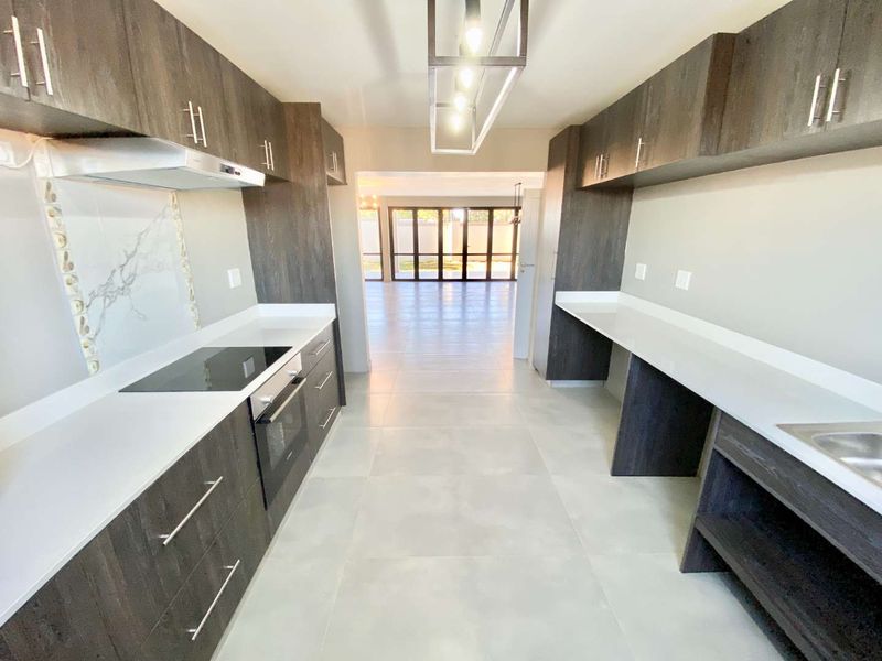 Modern galley-style kitchen featuring dark wood cabinetry, white quartz countertops, integrated appliances, and a stylish marble-look splashback, leading into an open-plan living area.