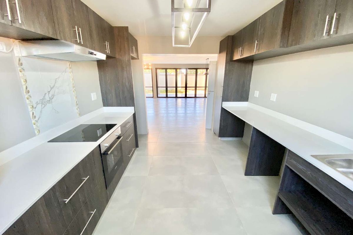 Modern galley-style kitchen featuring dark wood cabinetry, white quartz countertops, integrated appliances, and a stylish marble-look splashback, leading into an open-plan living area.