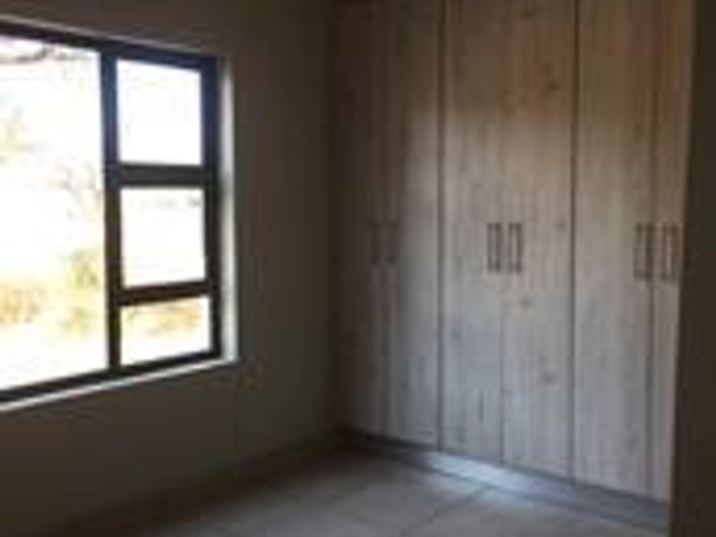 A clean, empty bedroom featuring modern light wood-finish built-in cupboards, neutral tiled flooring, and an aluminium-framed window.